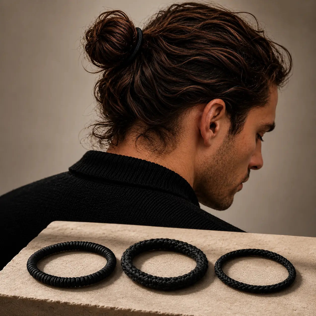 Man with styled hair sitting behind three black silk hair ties on a neutral background