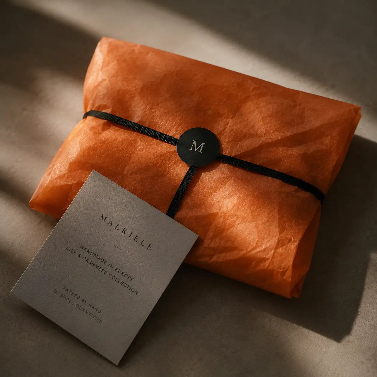 
                  
                    Gift wrapped in orange paper with a black ribbon and 'Malkiele' card on a textured surface.
                  
                