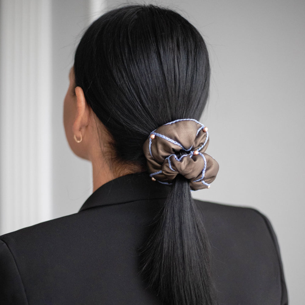 A black-haired woman, with stylish ponytail is wearing Malkiele designer brown vegan leather scrunchie, embellished with 6 pearls from Swarovski and light blue silk knit ribbon, name Ambitious.