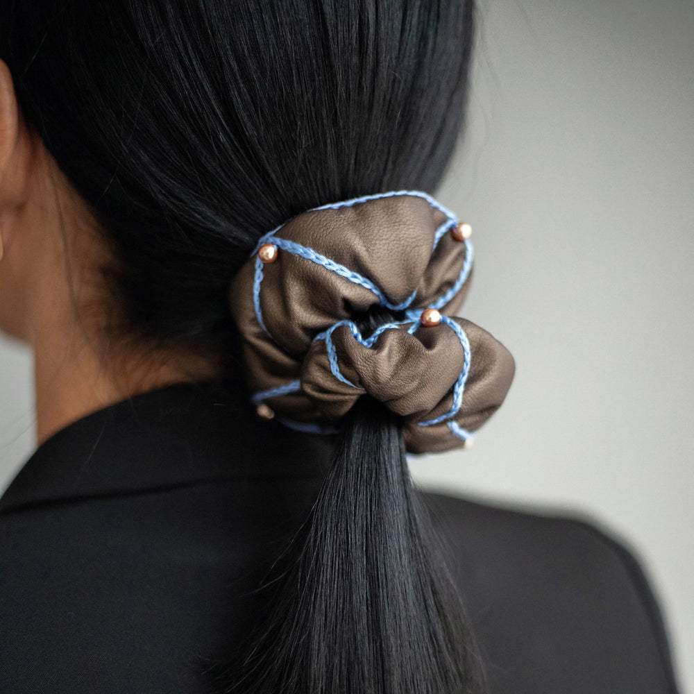 
                  
                    A black-haired woman, with stylish ponytail is wearing Malkiele designer brown vegan leather scrunchie, embellished with 6 pearls from Swarovski and light blue silk knit ribbon, name Ambitious.
                  
                