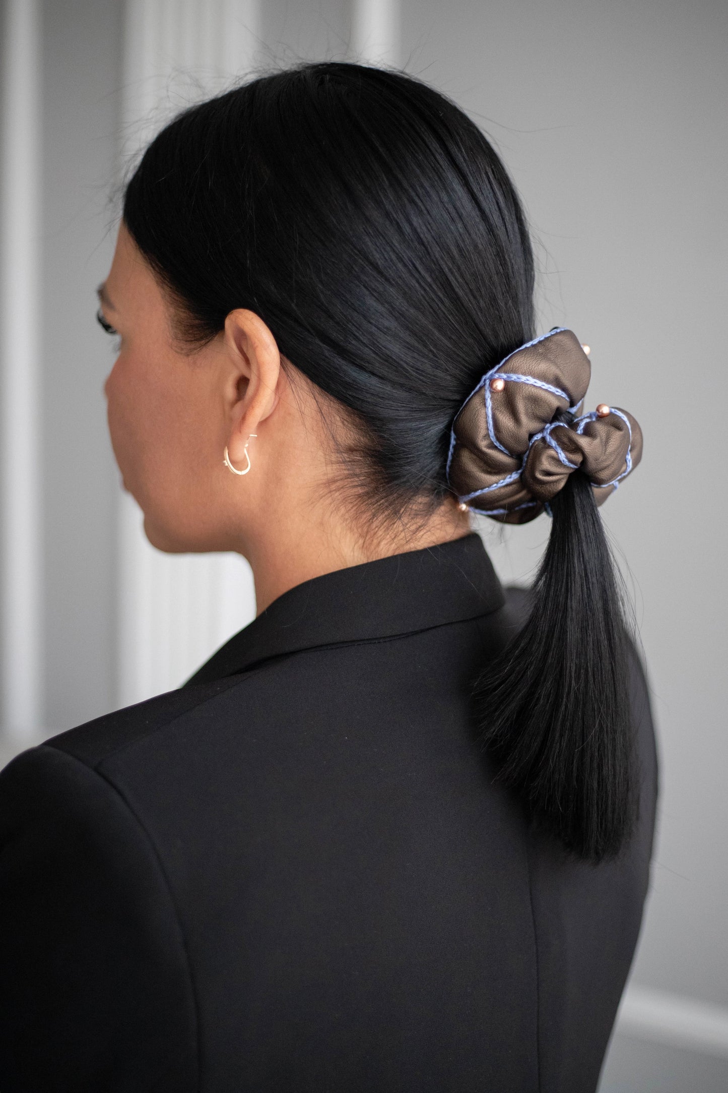 
                  
                    A black-haired woman, with stylish ponytail is wearing Malkiele designer brown vegan leather scrunchie, embellished with 6 pearls from Swarovski and light blue silk knit ribbon, name Ambitious.
                  
                