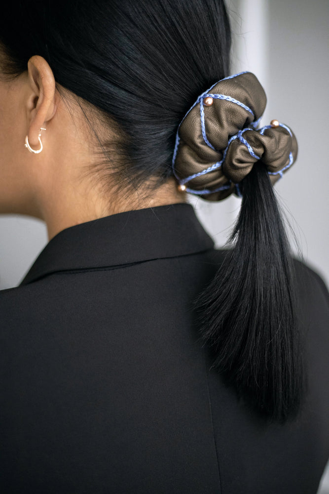 
                  
                    A black-haired woman, with stylish ponytail is wearing Malkiele designer brown vegan leather scrunchie, embellished with 6 pearls from Swarovski and light blue silk knit ribbon, side view, name Ambitious.
                  
                