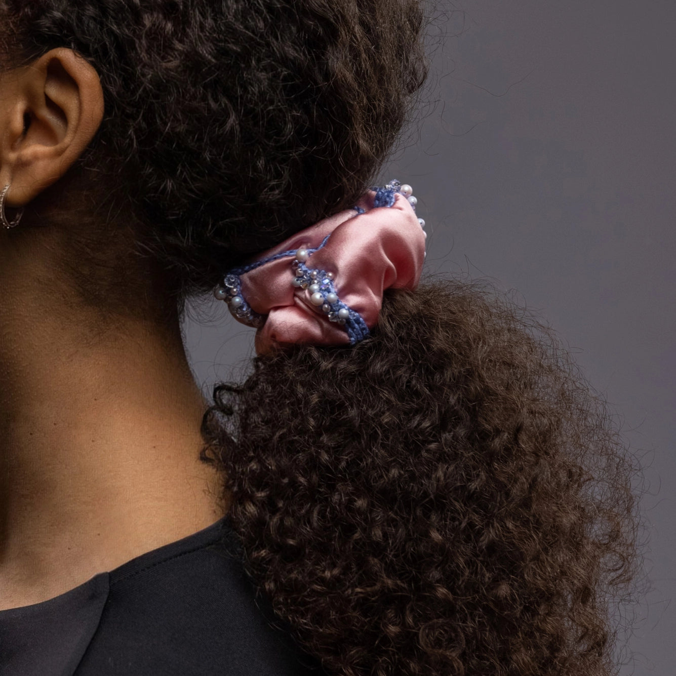 
                  
                    A dark-haired girl, details, with stylish ponytail is wearing Malkiele designer pink silk scrunchie, embellished with crystals from Swarovski and light blue silk knit ribbon, name Elizabeth.
                  
                