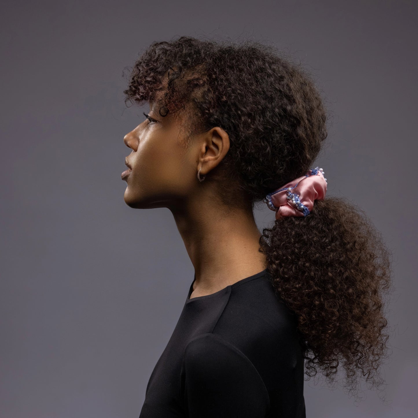 A dark-haired girl, side view, with stylish ponytail is wearing Malkiele designer pink silk scrunchie, embellished with crystals from Swarovski and light blue silk knit ribbon, name Elizabeth.