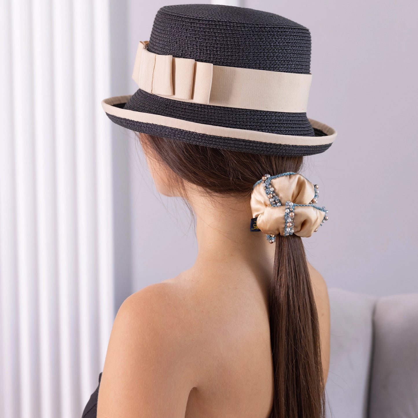 
                  
                    A dark-haired girl in hat, view from the back, with stylish ponytail is wearing Malkiele designer beige silk scrunchie, embellished with crystals from Swarovski® and green silk knit ribbon, name Elizabeth.
                  
                