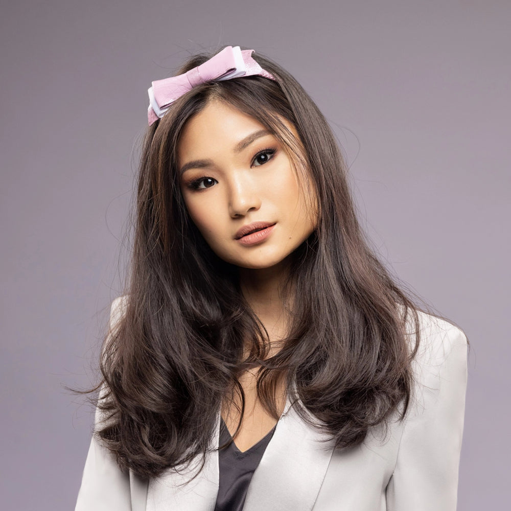 A dark-haired asian woman is wearing Malkiele designer pink silk knit headbands, embellished with pink and silver leather bow, name Enjoyable.