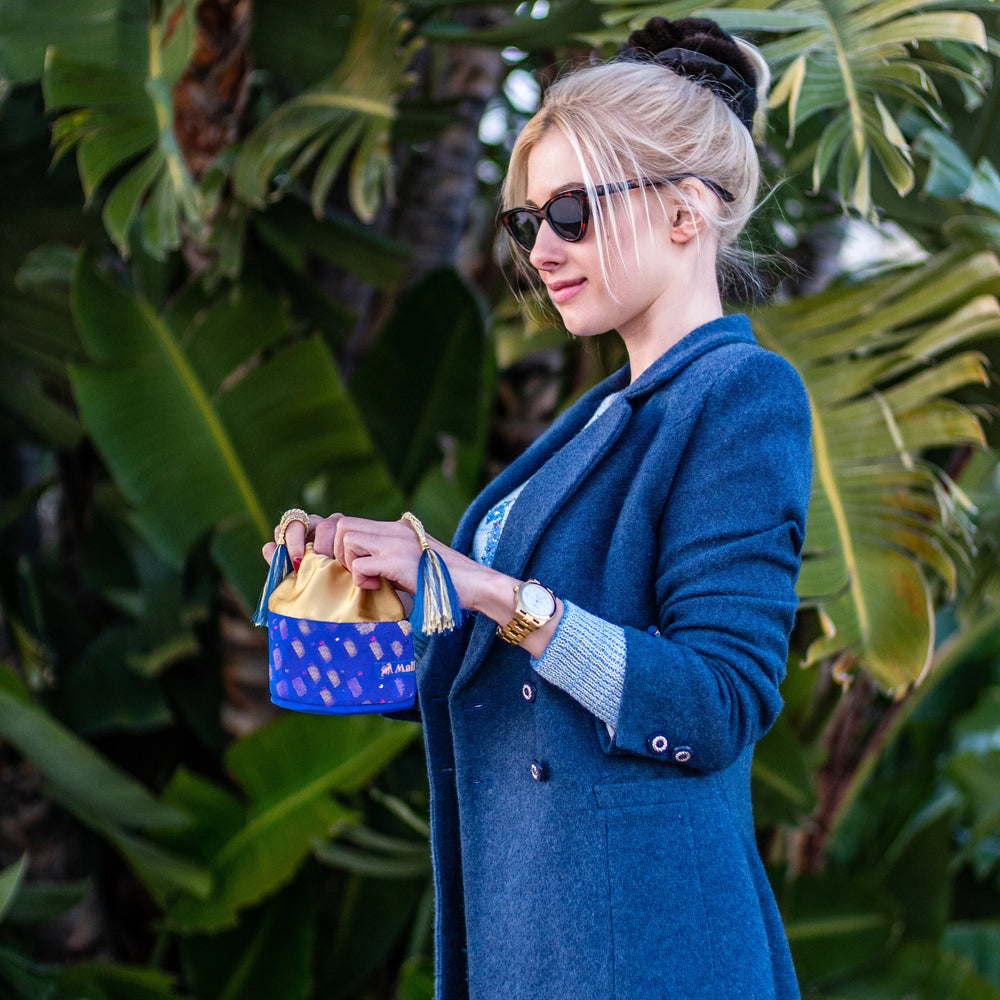 
                  
                    Blonde-haired girl in blue jacket is opening royal blue round art suede bag, with gold silk lining, blue tassels, Malkiele logo, embellished with crystals from Swarovski®, name Favourite.
                  
                