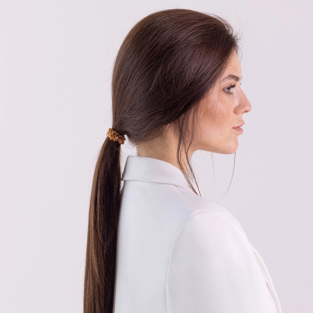 
                  
                    A dark-haired girl in white jacket, side view with half face, with ponytail is wearing Malkiele designer beige silk gentle hair tie, name Finesse.
                  
                