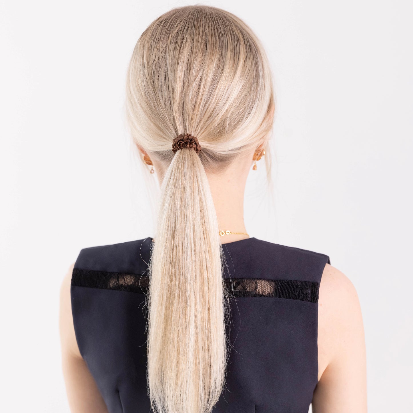 
                  
                    A blonde-haired girl in black dress, view from the back, with ponytail is wearing Malkiele designer brown silk gentle hair tie, name Finesse.
                  
                