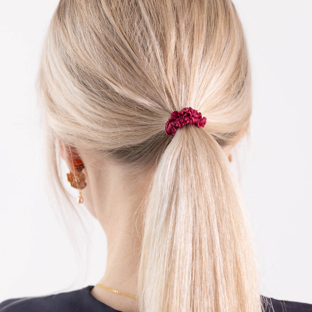 
                  
                    A blonde-haired girl in black dress, view from the back, with ponytail is wearing Malkiele designer burgundy silk gentle hair tie, name Finesse.
                  
                