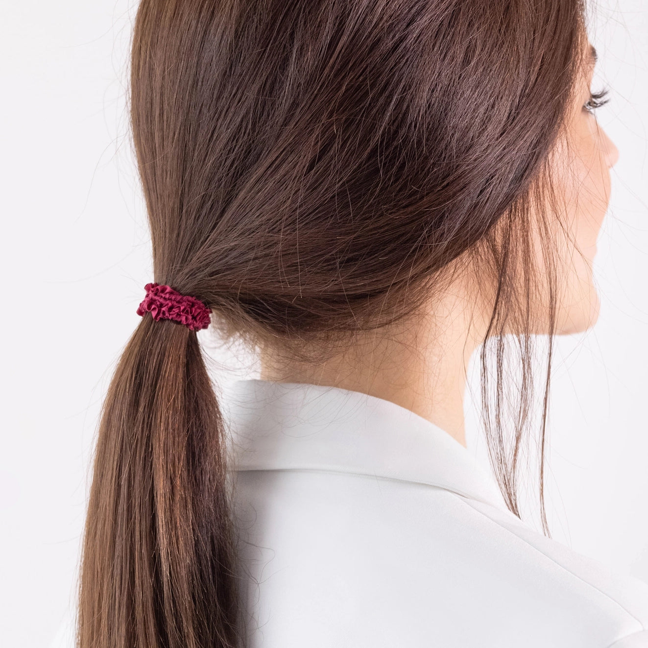 
                  
                    A dark-haired girl in white jacket, side view, with ponytail is wearing Malkiele designer burgundy silk gentle hair tie, name Finesse.
                  
                