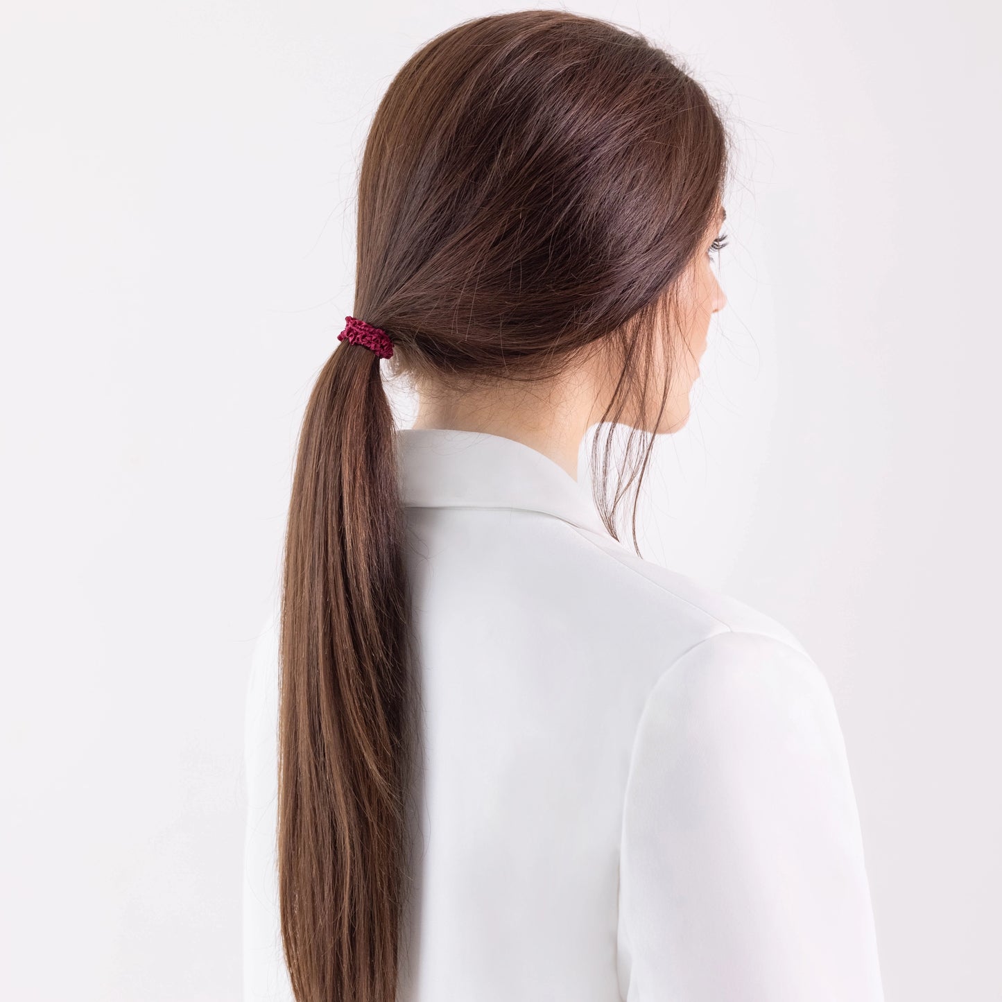 A dark-haired girl in white jacket with ponytail is wearing Malkiele designer burgundy silk gentle hair tie, name Finesse.