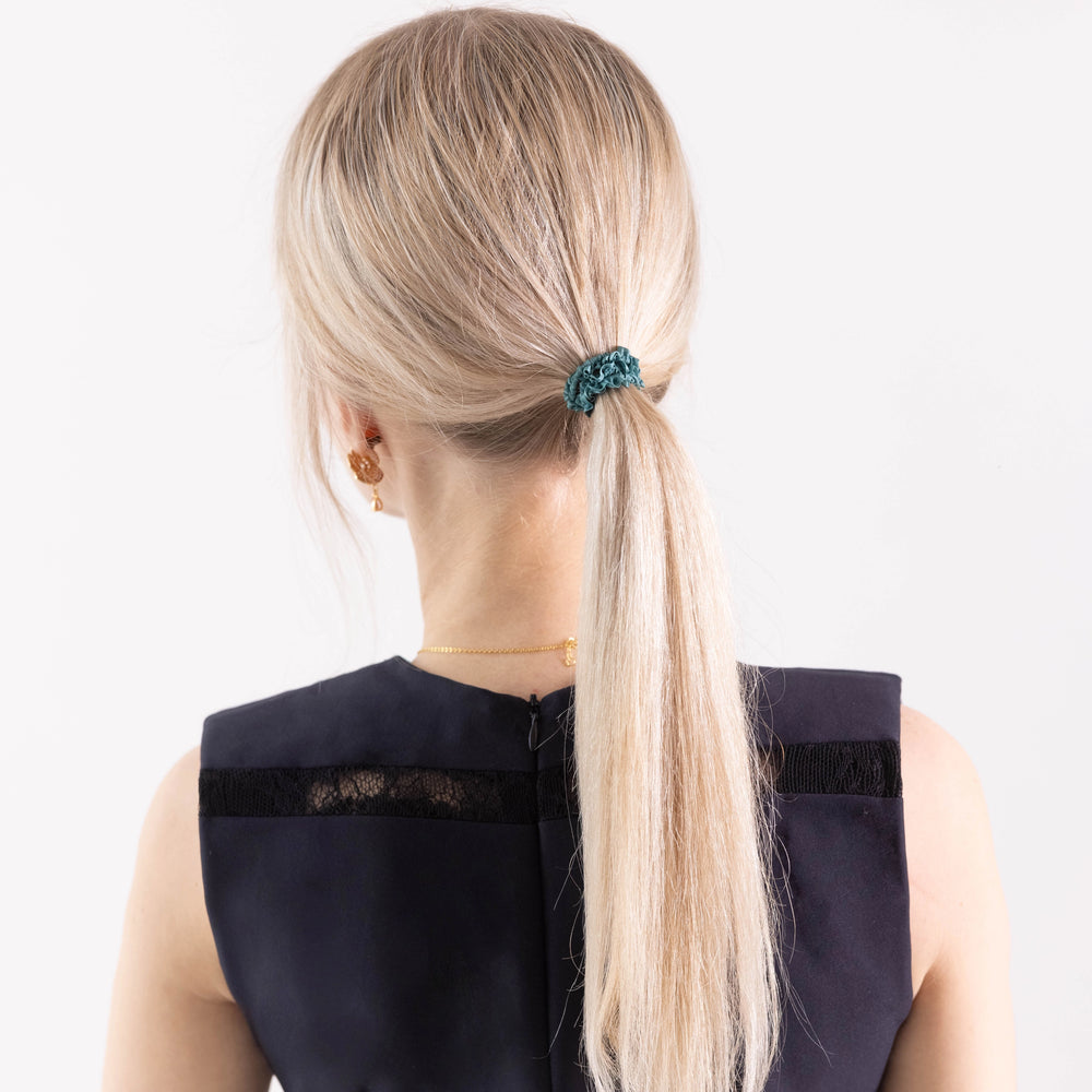 
                  
                    A blonde-haired girl in black dress, view from the back, with ponytail is wearing Malkiele designer green silk gentle hair tie, name Finesse.
                  
                