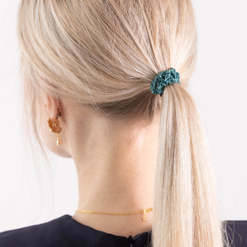 
                  
                    A blonde-haired girl in black dress, view from the back, details, with ponytail is wearing Malkiele designer green silk gentle hair tie, name Finesse.
                  
                