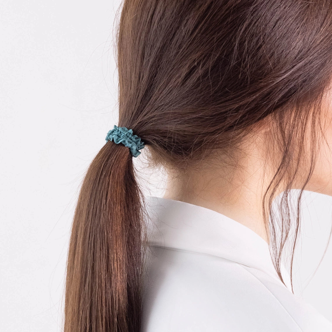 
                  
                    A dark-haired girl in white jacket, details view, with ponytail is wearing Malkiele designer green silk gentle hair tie, name Finesse.
                  
                