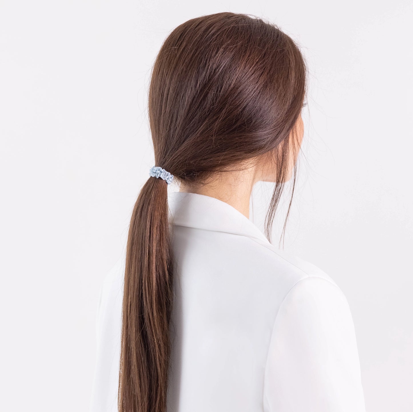 
                  
                    A dark-haired girl in white jacket, view from the back, with classic ponytail is wearing Malkiele designer grey silk gentle hair tie, name Finesse.
                  
                