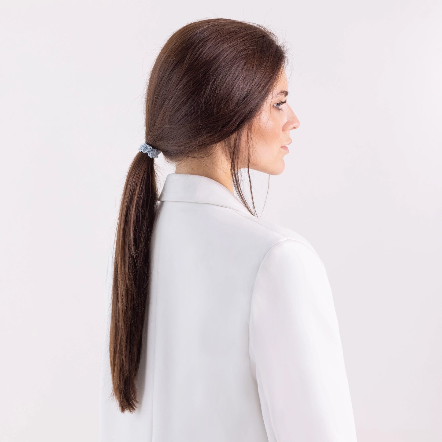A dark-haired girl in white jacket, side view with half face, with classic ponytail is wearing Malkiele designer grey silk gentle hair tie, name Finesse.