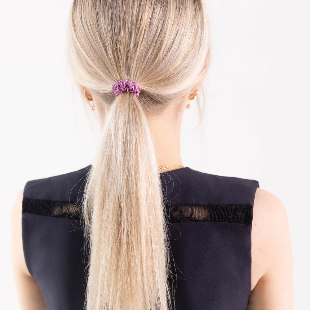 
                  
                    A blonde-haired girl in black dress, view from the back, with classic ponytail is wearing Malkiele designer pink silk gentle hair tie, name Finesse.
                  
                