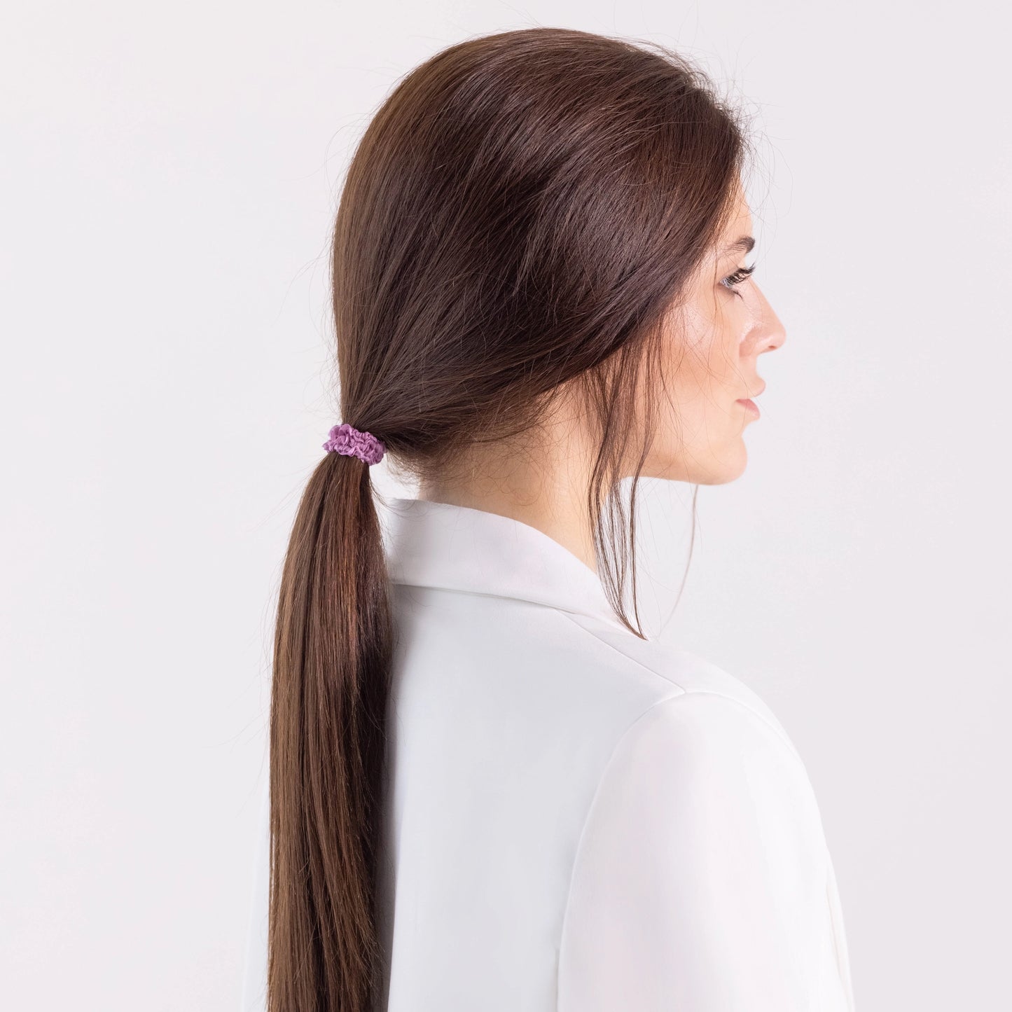 A dark-haired girl in white jacket, side view with half face, with classic ponytail is wearing Malkiele designer pink silk gentle hair tie, name Finesse.