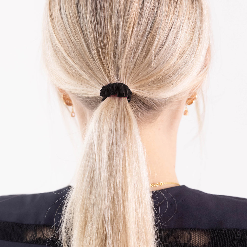 
                  
                    A blonde-haired girl in black dress, view from the back, with classic ponytail is wearing Malkiele designer black silk gentle hair tie, name Finesse.
                  
                