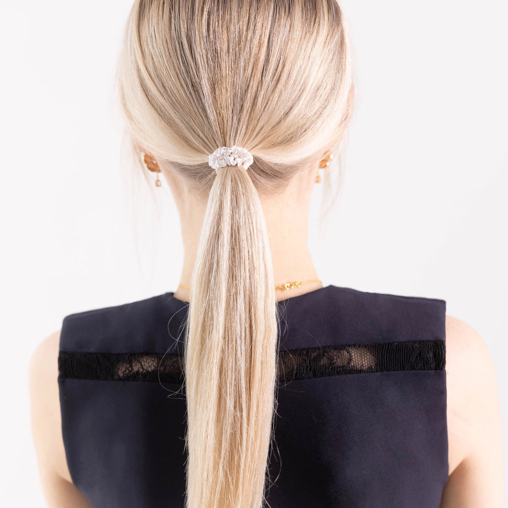 
                  
                    A blonde-haired girl in black dress, view from the back, with classic ponytail is wearing Malkiele designer white silk gentle hair tie, name Finesse.
                  
                