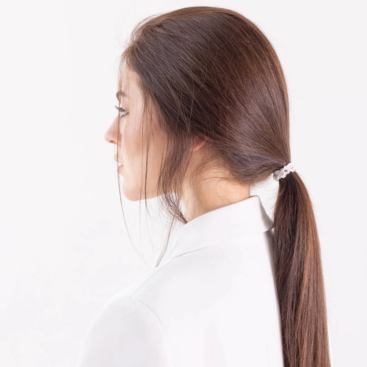 
                  
                    A dark-haired girl in white jacket, side view with half face, details, with classic ponytail is wearing Malkiele designer bridal white silk gentle hair tie, name Finesse.
                  
                