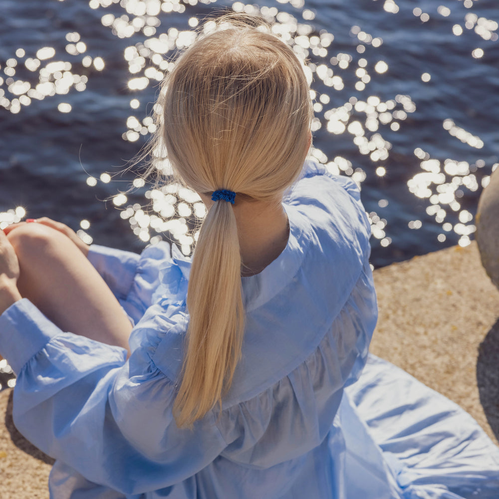 A blonde-haired girl in light blue dress, from behind with classic ponytail is wearing Malkiele designer royal blue silk hair tie, name Finesse.