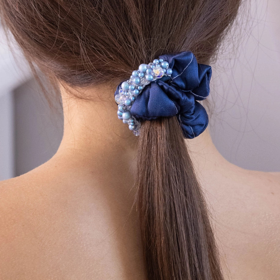 
                  
                    A dark-haired woman, view from the back, scrunchie details, with stylish ponytail is wearing Malkiele designer navy blue silk scrunchie, embellished with light blue pearls from Swarovski® and grey silk knit ribbon, name Glowing.
                  
                