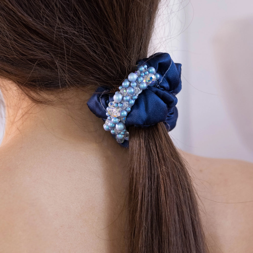 
                  
                    A dark-haired woman, view from the back, more scrunchie details, with stylish ponytail is wearing Malkiele designer navy blue silk scrunchie, embellished with light blue pearls from Swarovski® and grey silk knit ribbon, name Glowing.
                  
                