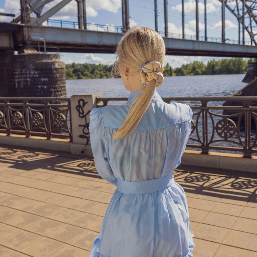 
                  
                    A blonde-haired girl in light blue dress, from behind, with stylish ponytail is wearing Malkiele designer beige silk scrunchie, embellished with 6 white pearls from Swarovski and light blue silk knit ribbon, name Majestic.
                  
                