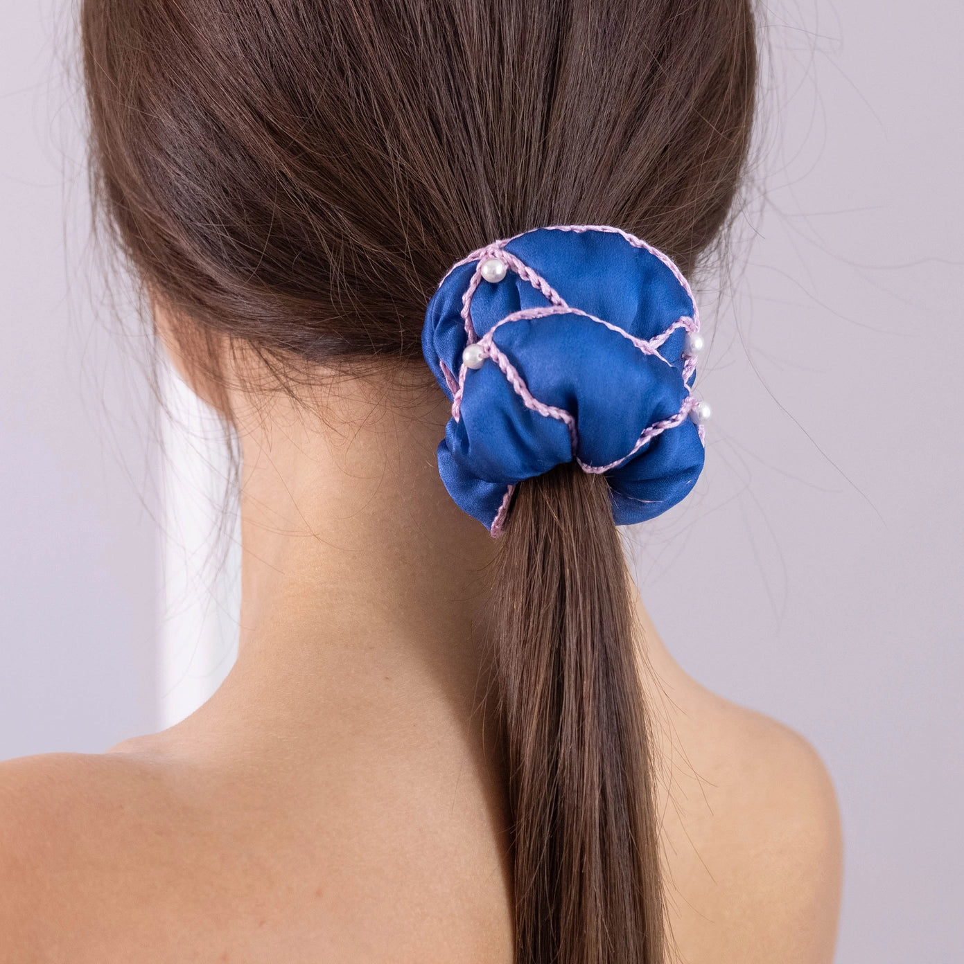 
                  
                    A dark-haired woman, view from the back, with stylish ponytail is wearing Malkiele designer royal blue silk scrunchie, embellished with 6 white pearls from Swarovski and pink silk knit ribbon, name Majestic.
                  
                