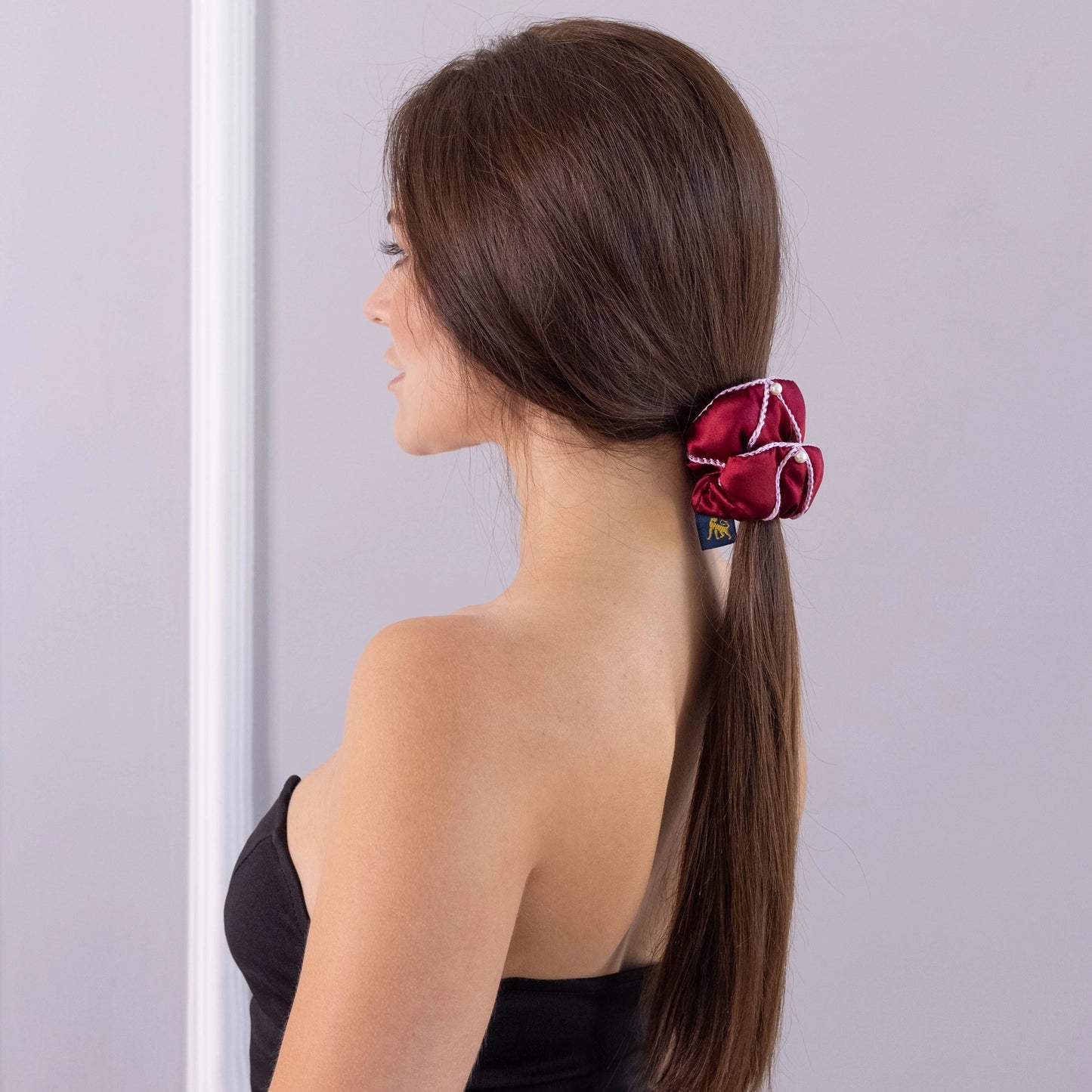 A dark-haired woman, view from the back, with stylish ponytail is wearing Malkiele designer burgundy silk scrunchie, embellished with 6 white pearls from Swarovski and pink silk knit ribbon, name Majestic.