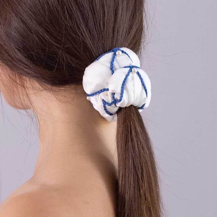 
                  
                    A dark-haired woman, view from the back, with stylish ponytail is wearing Malkiele designer white silk scrunchie, embellished with 6 white pearls from Swarovski and blue silk knit ribbon, name Majestic.
                  
                