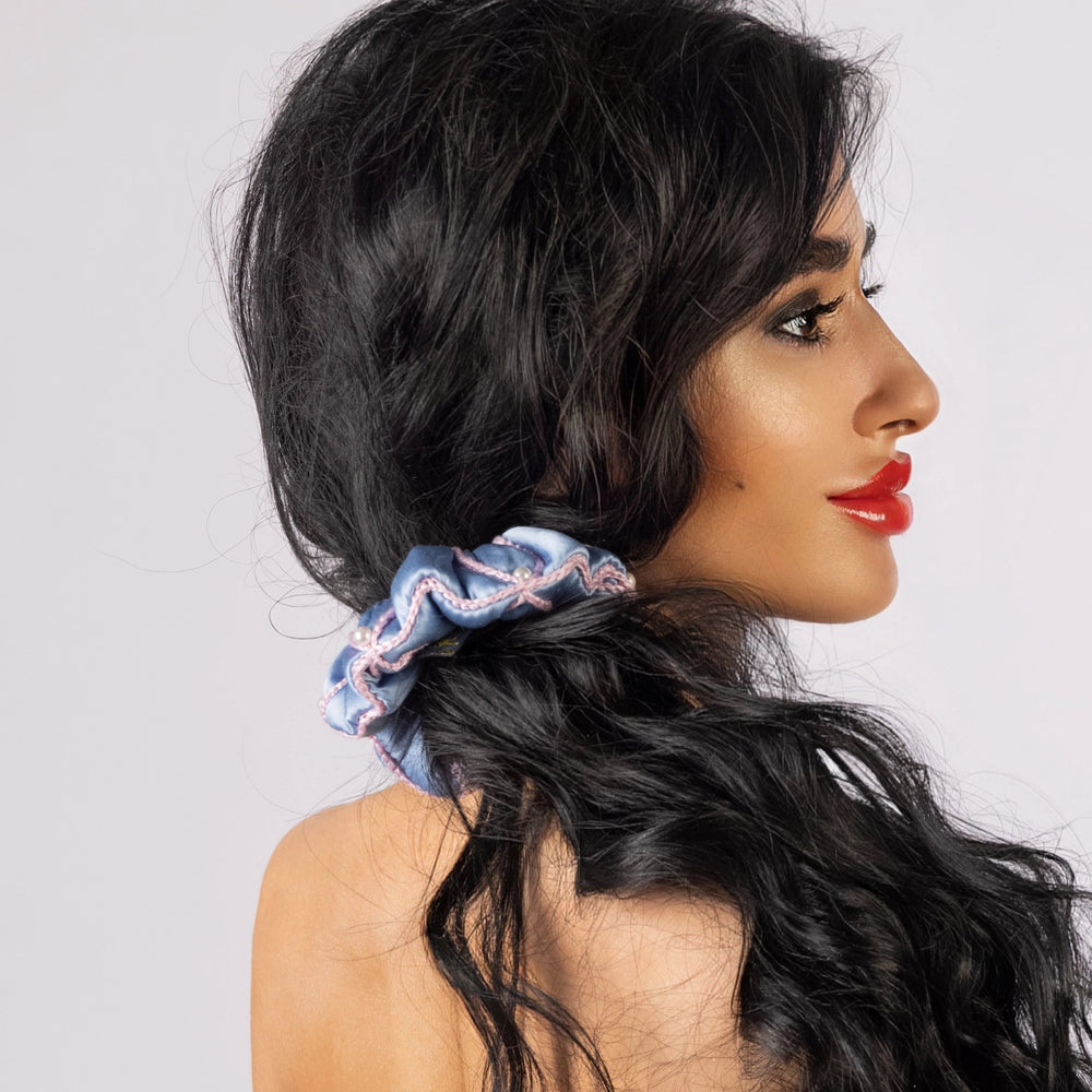 
                  
                    A black-haired woman, with stylish ponytail is wearing Malkiele designer light blue silk scrunchie, embellished with 6 white pearls from Swarovski and pink silk knit ribbon, name Majestic.
                  
                