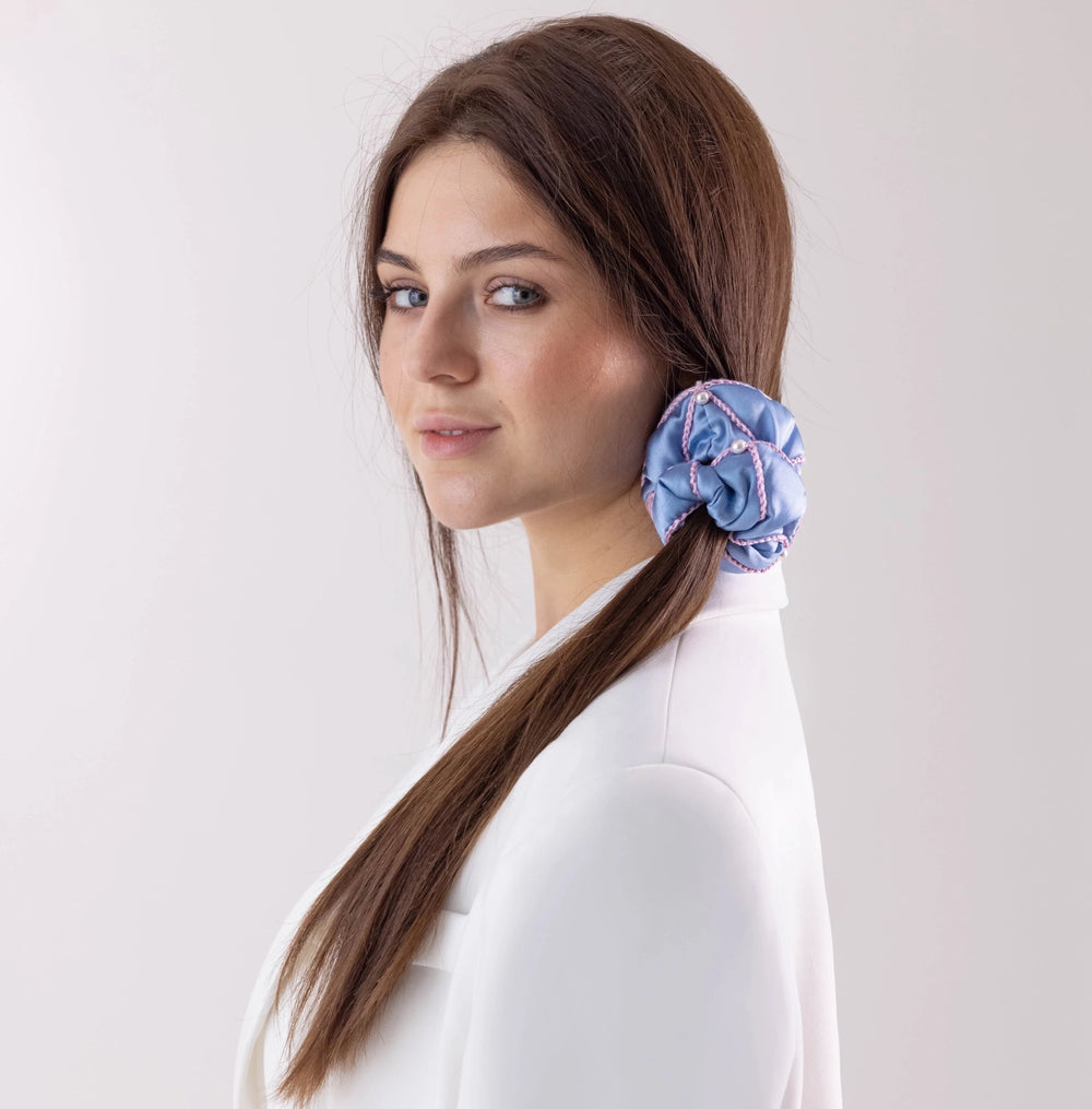 A dark-haired woman in white jacket, with stylish ponytail is wearing Malkiele designer light blue silk scrunchie, embellished with 6 white pearls from Swarovski® and pink silk knit ribbon, name Majestic.