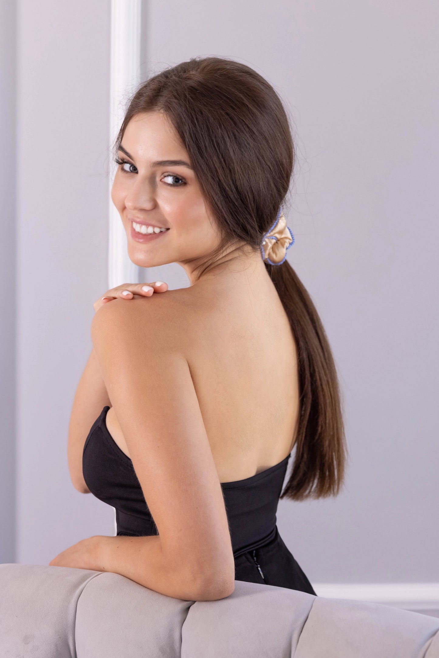 
                  
                    A dark-haired smiling woman, with stylish ponytail is wearing Malkiele designer beige silk scrunchie, embellished with 6 white pearls from Swarovski and light blue silk knit ribbon, name Majestic.
                  
                