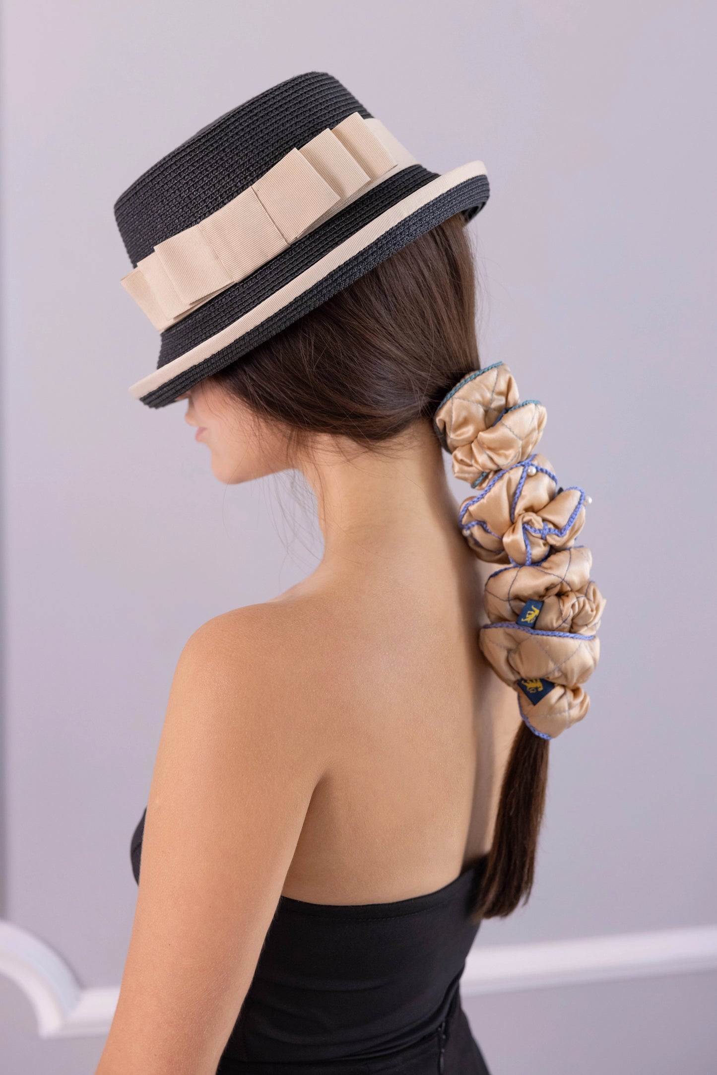 
                  
                    A dark-haired woman in hat, side view, with stylish ponytail is wearing Malkiele designer beige silk scrunchies set, embellished with 6 white pearls from Swarovski and light blue silk knit ribbon, name Majestic.
                  
                