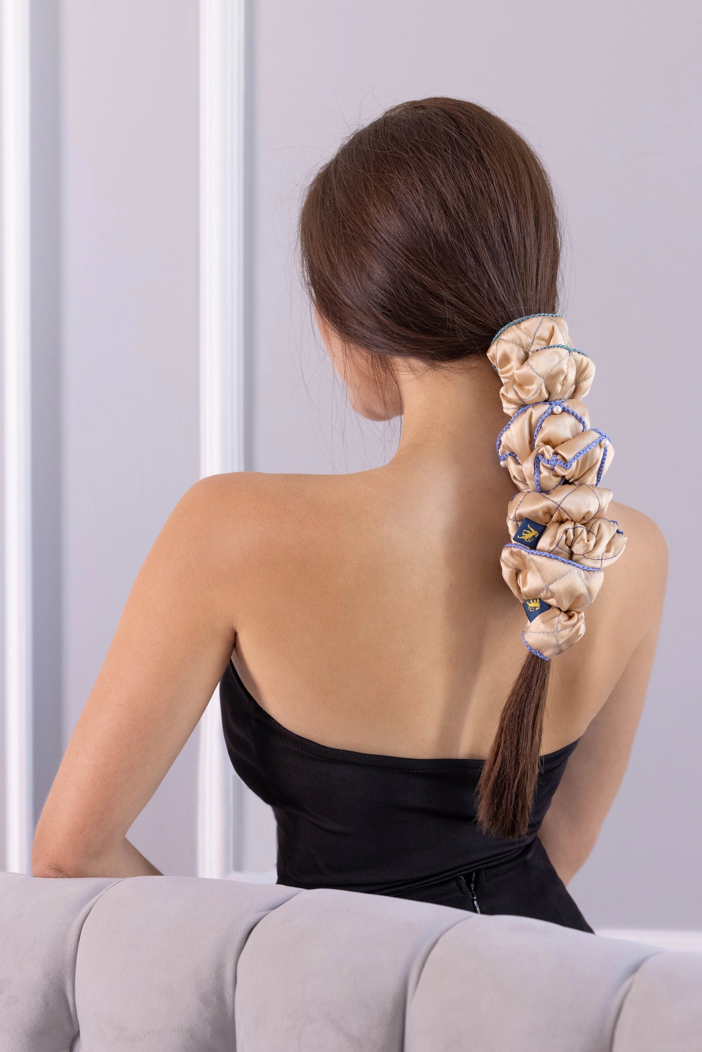 
                  
                    A dark-haired woman, view from the back, with stylish ponytail is wearing Malkiele designer beige silk scrunchies set, embellished with 6 white pearls from Swarovski and light blue silk knit ribbon, name Majestic.
                  
                