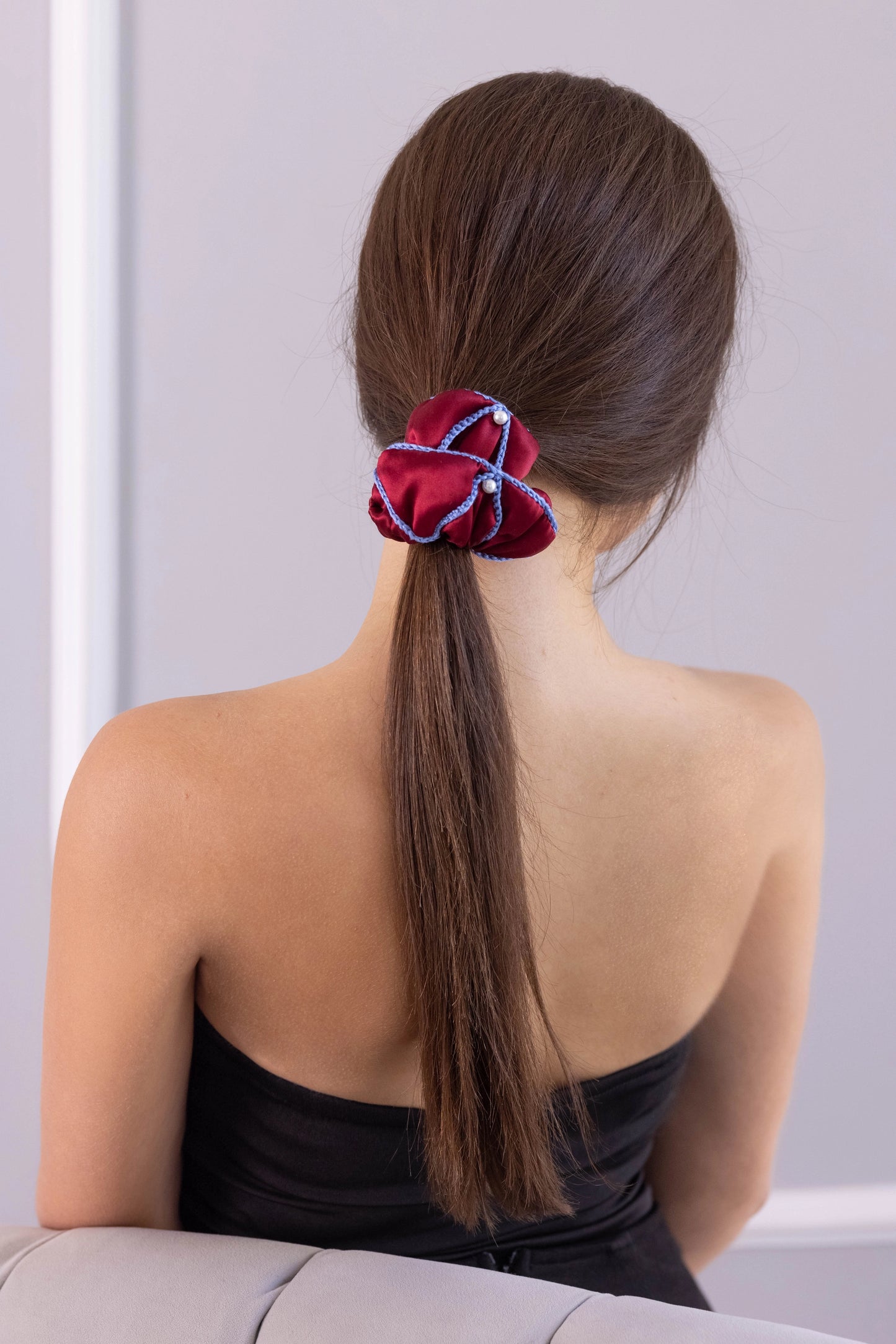 
                  
                    A dark-haired woman, view from the back, with stylish ponytail is wearing Malkiele designer burgundy silk scrunchie, embellished with 6 white pearls from Swarovski and light blue silk knit ribbon, name Majestic.
                  
                