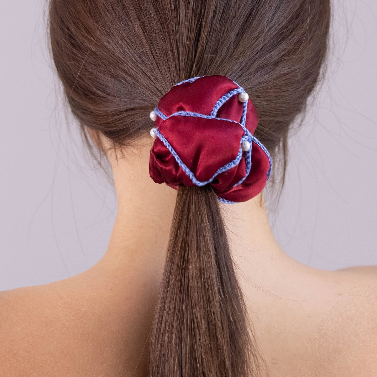 
                  
                    A dark-haired woman, view from the back, with stylish ponytail is wearing Malkiele designer burgundy silk scrunchie, embellished with 6 white pearls from Swarovski and light blue silk knit ribbon, name Majestic.
                  
                