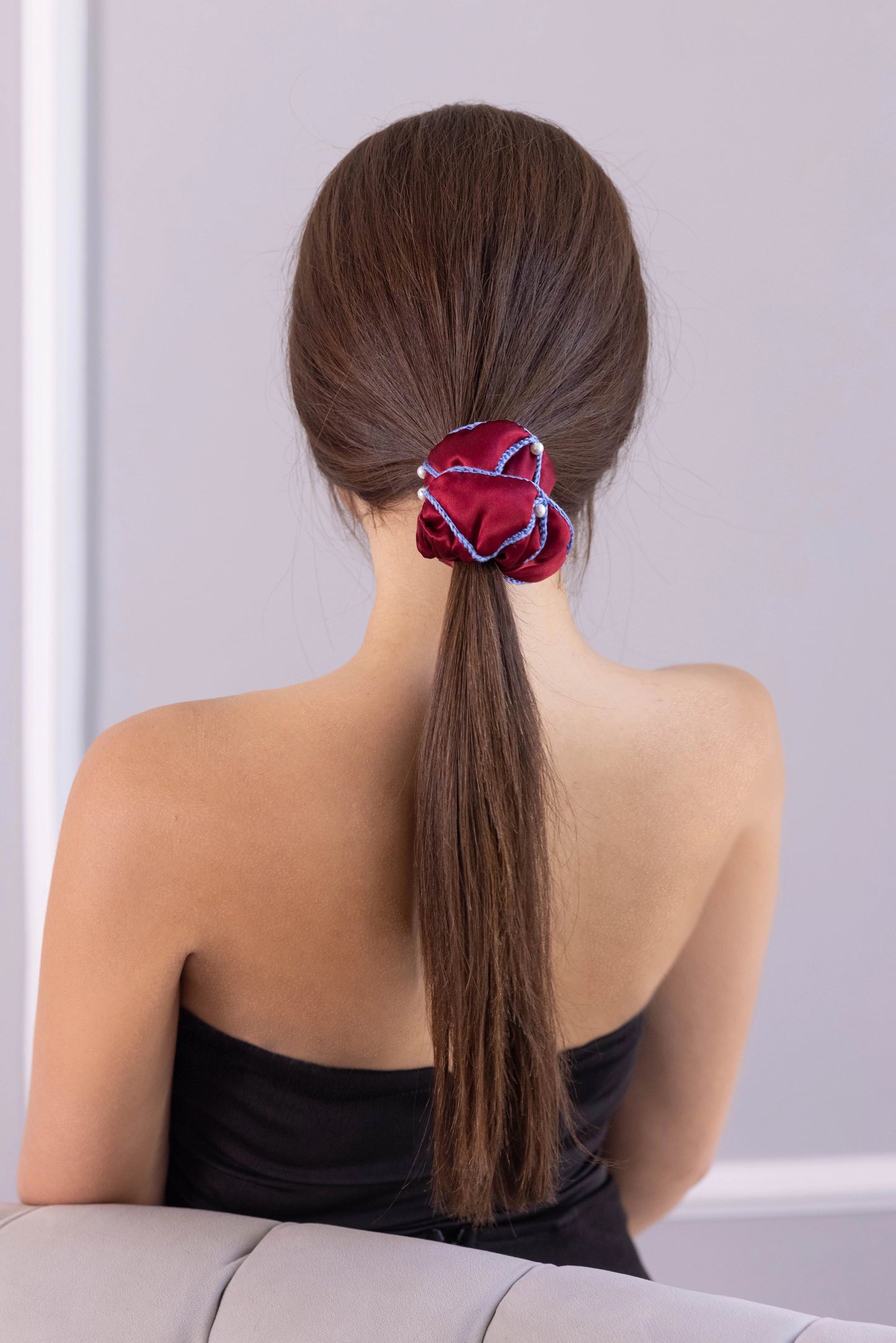 
                  
                    A dark-haired woman, view from the back, with stylish ponytail is wearing Malkiele designer burgundy silk scrunchie, embellished with 6 white pearls from Swarovski and light blue silk knit ribbon, name Majestic.
                  
                