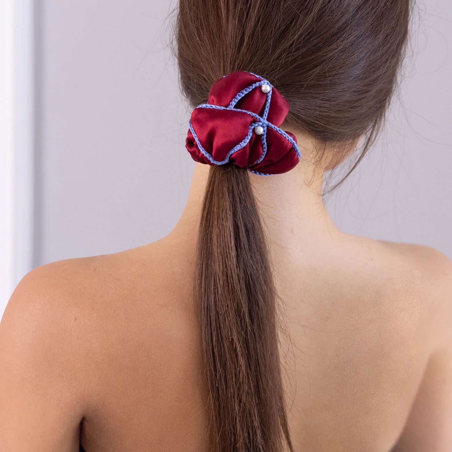 
                  
                    A dark-haired woman, view from the back, with stylish ponytail is wearing Malkiele designer burgundy silk scrunchie, embellished with 6 white pearls from Swarovski and light blue silk knit ribbon, name Majestic.
                  
                