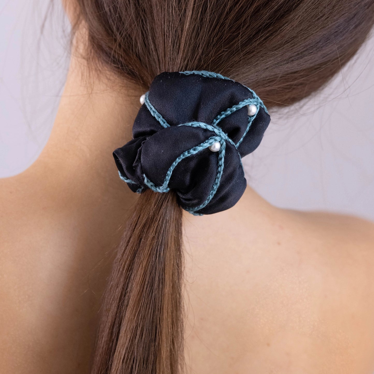 
                  
                    A dark-haired woman, with stylish ponytail is wearing Malkiele designer black silk scrunchie, embellished with 6 white pearls from Swarovski and green silk knit ribbon, name Majestic.
                  
                