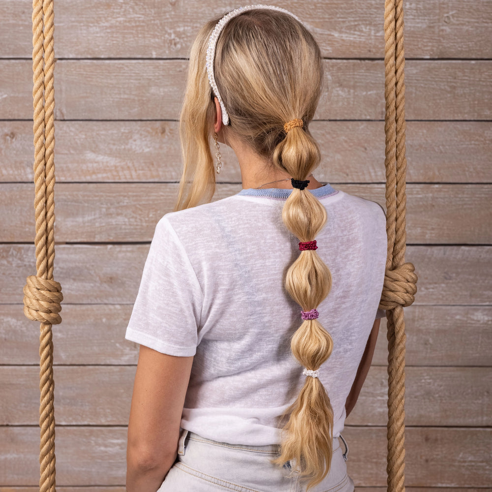 
                  
                    A blonde-haired girl in white t-shirt, view from the back, is sitting on a swing with ropes, her ponytail is wearing five Malkiele designer mixed colors silk gentle hair ties, name Finesse and white silk headband.
                  
                