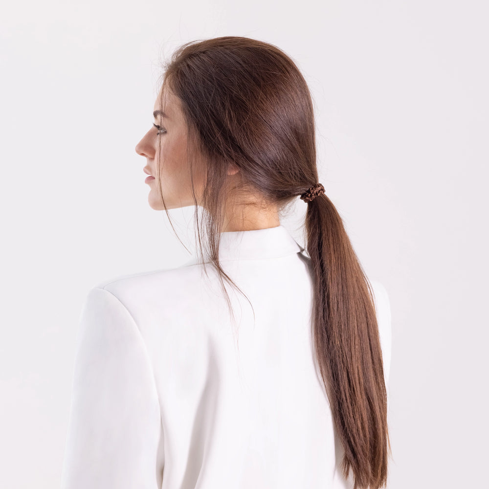 
                  
                    A dark-haired girl in white jacket, side view with half face, with a stylish ponytail is wearing Malkiele designer brown silk hair tie, name Finesse.
                  
                