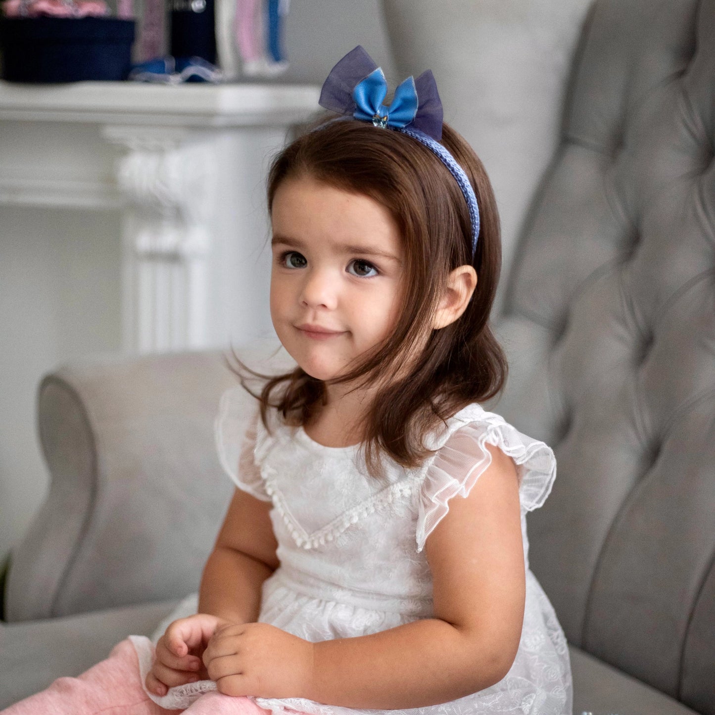 
                  
                    Dark-haired little girl in white dress is sitting on the sofa and is wearing light blue silk knit headband, embellished with crystal from Swarovski® and leather bow, Malkiele designer, name Miss Marie.
                  
                