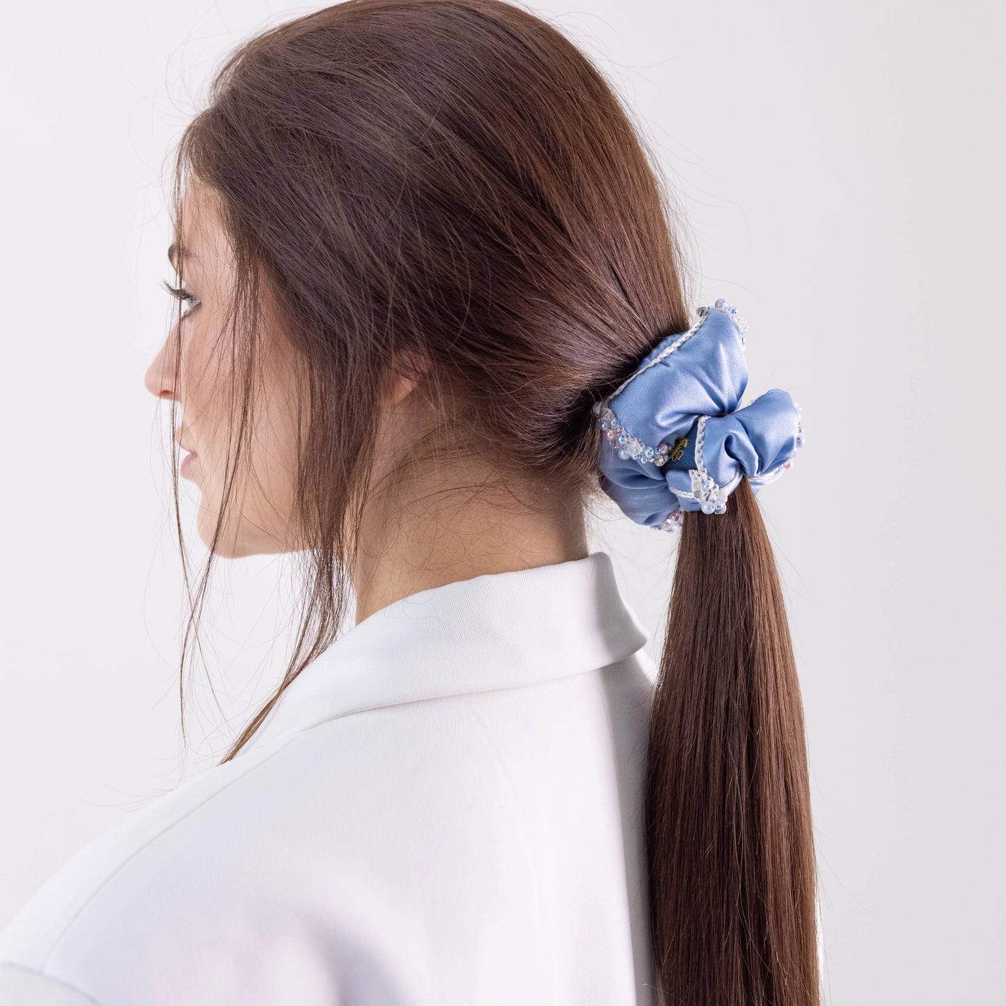 Dark-haired girl in white jacket, with stylish ponytail is wearing Malkiele designer light blue silk scrunchie, embellished with crystals from Swarovski® and white silk knit ribbon, name Elizabeth.