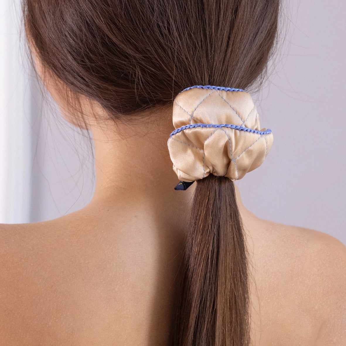 
                  
                    A dark-haired woman, view from the back, with stylish ponytail is wearing Malkiele designer beige silk scrunchie, embellished light blue silk knit ribbon, name Morning Passion.
                  
                