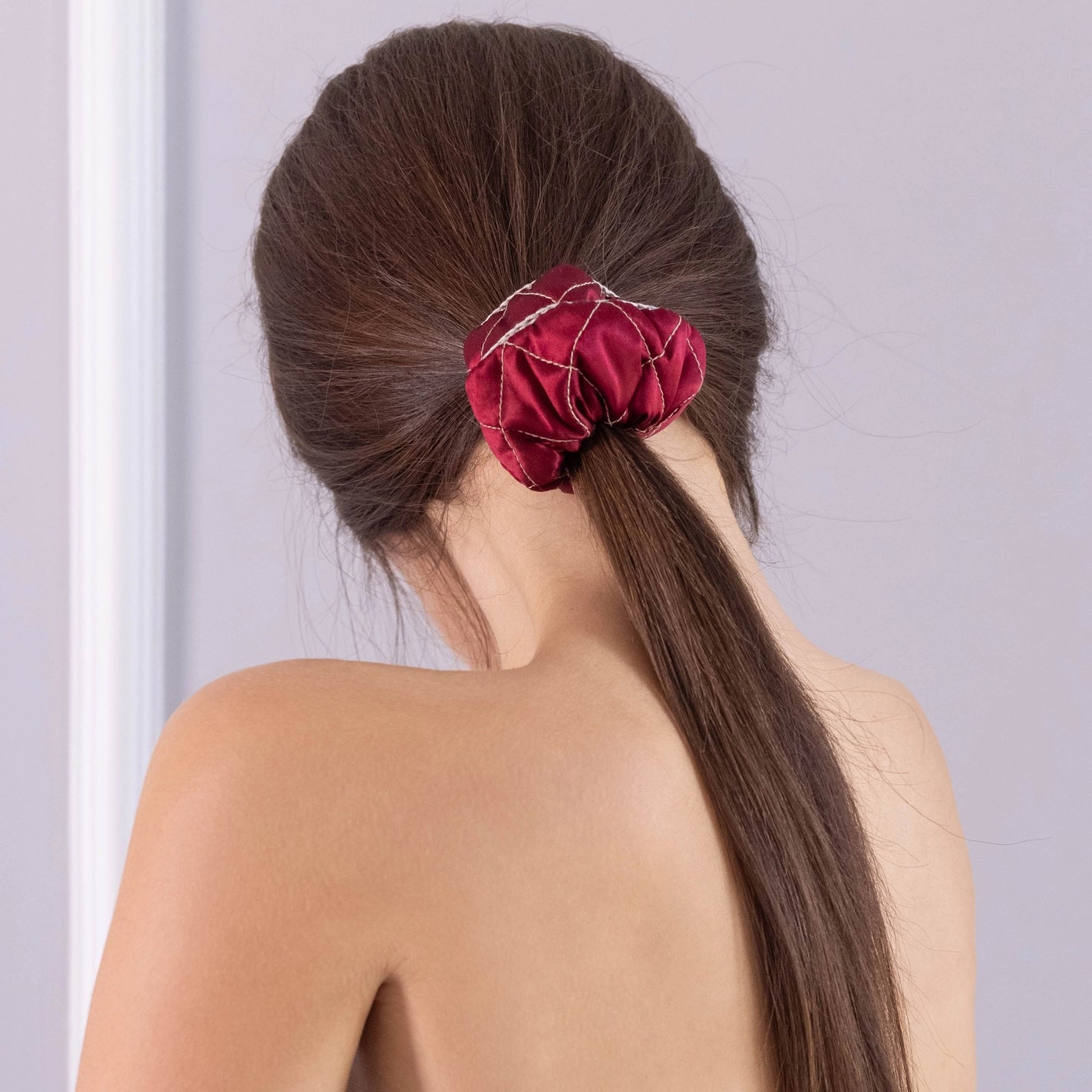 
                  
                    A dark-haired woman, view from the back, with stylish ponytail is wearing Malkiele designer burgundy silk scrunchie, embellished beige silk knit ribbon, name Morning Passion.
                  
                