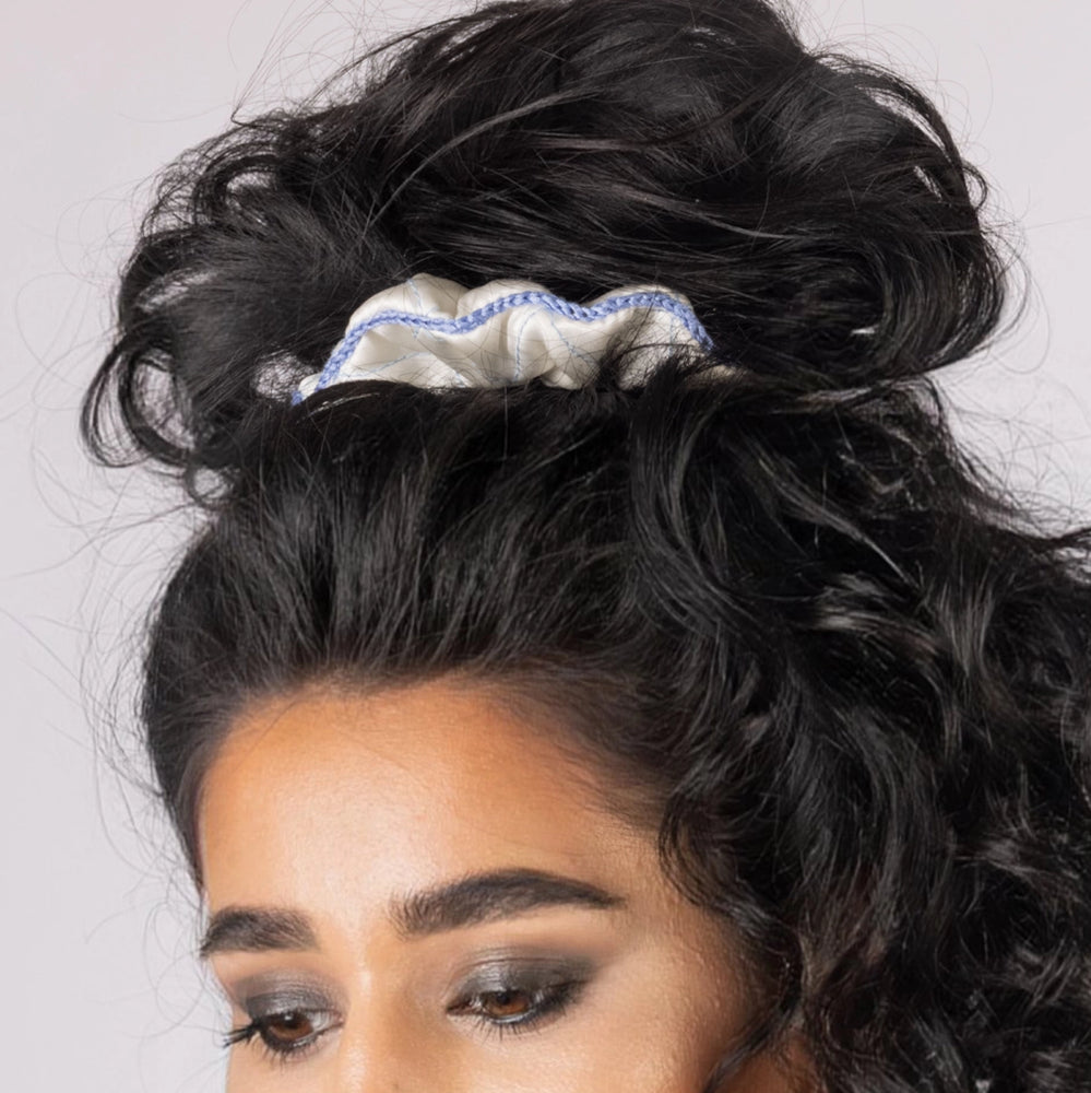 
                  
                    A dark-haired smiling woman, with stylish ponytail is wearing Malkiele designer white silk scrunchie, embellished light blue silk knit ribbon, name Morning Passion.
                  
                